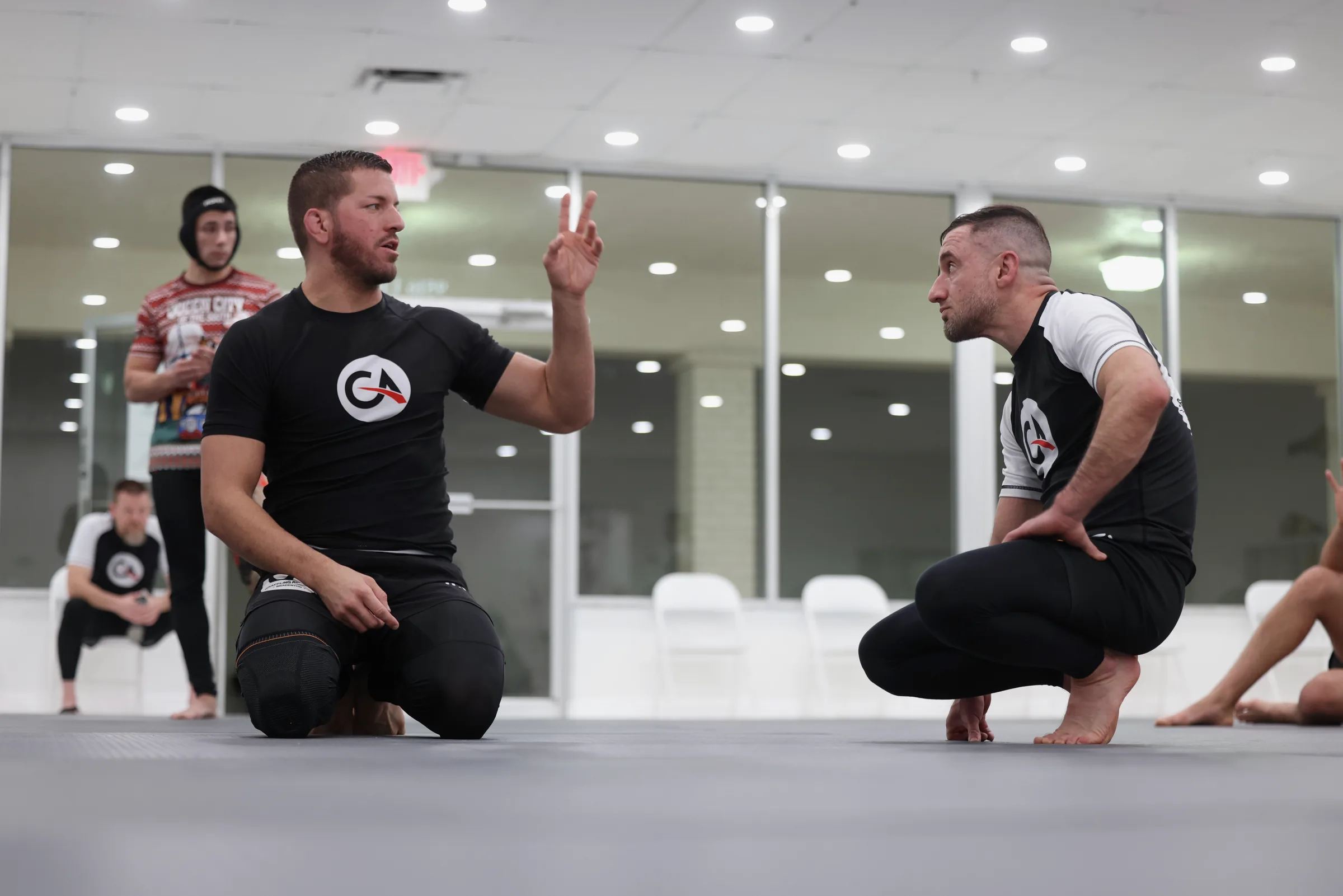 Two jiu-jitsu practitioners kneeling on a grey mat in conversation. One gestures with a raised hand while speaking; the other squats in an attentive listening posture. Other training partners visible in the background. Bright institutional lighting reflects in the mirrored wall behind them.